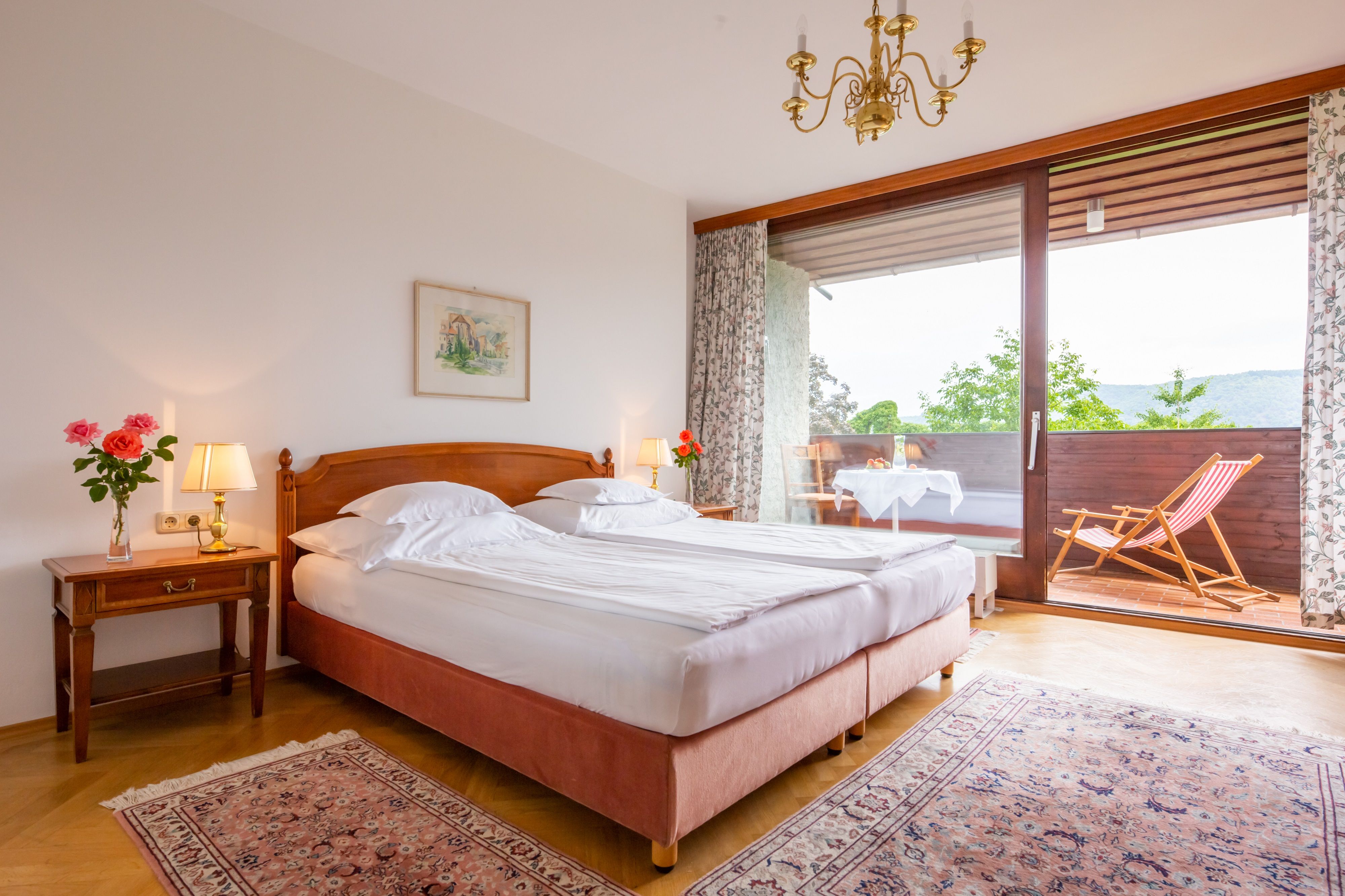 Ein gemütliches Schlafzimmer mit Doppelbett, Nachttischlampen und Balkon mit Liegestuhl und Tisch.