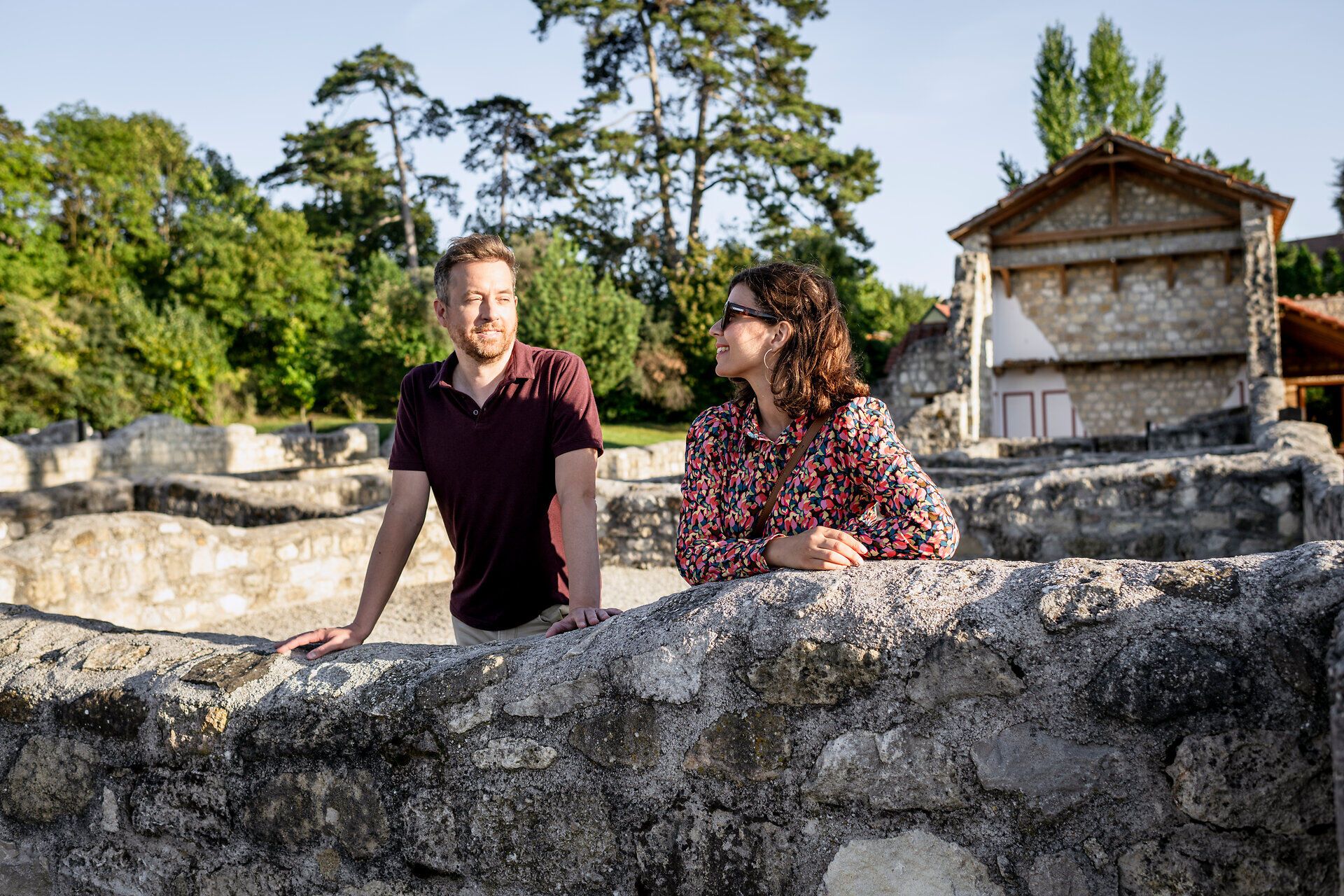Paar lehnt an einer Steinmauer im Römischen Stadtviertel der Römerstadt Caruntum, im Hintergrund ein Steinhaus mit Wandmalerei. 