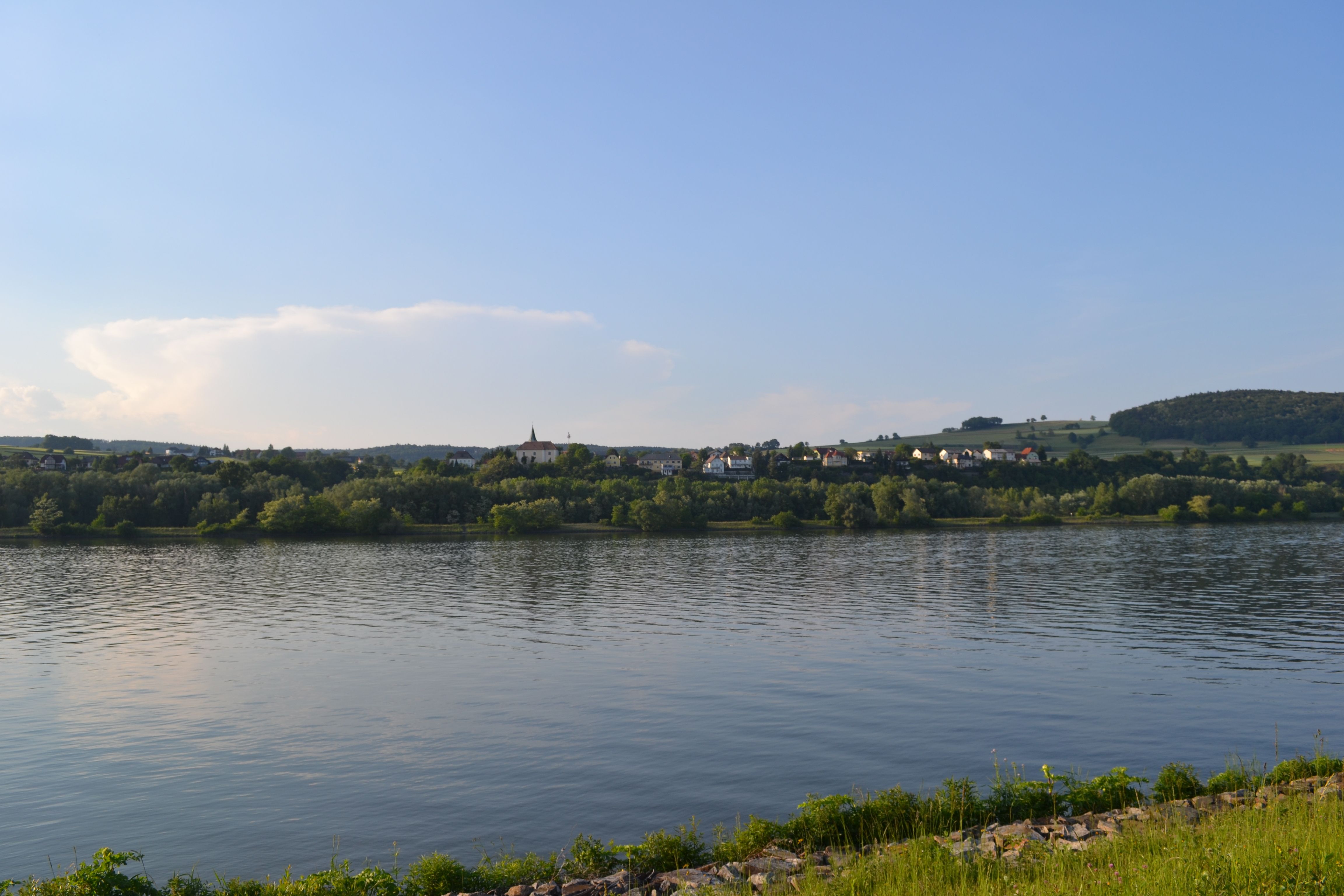 Landschaft mit Fluss, Hügeln und Dorf im Hintergrund bei klarem Himmel.