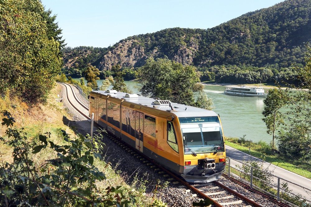 A yellow train of the Wachau Railway runs along a river, with a cruise ship and wooded hills in the background.