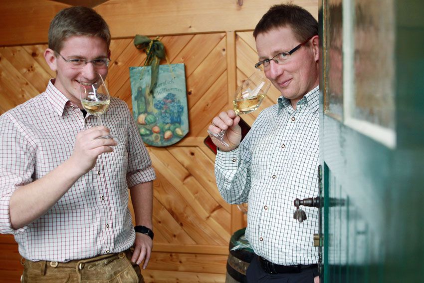 Zwei Männer in karierten Hemden trinken Weißwein in einem holzgetäfelten Raum.
