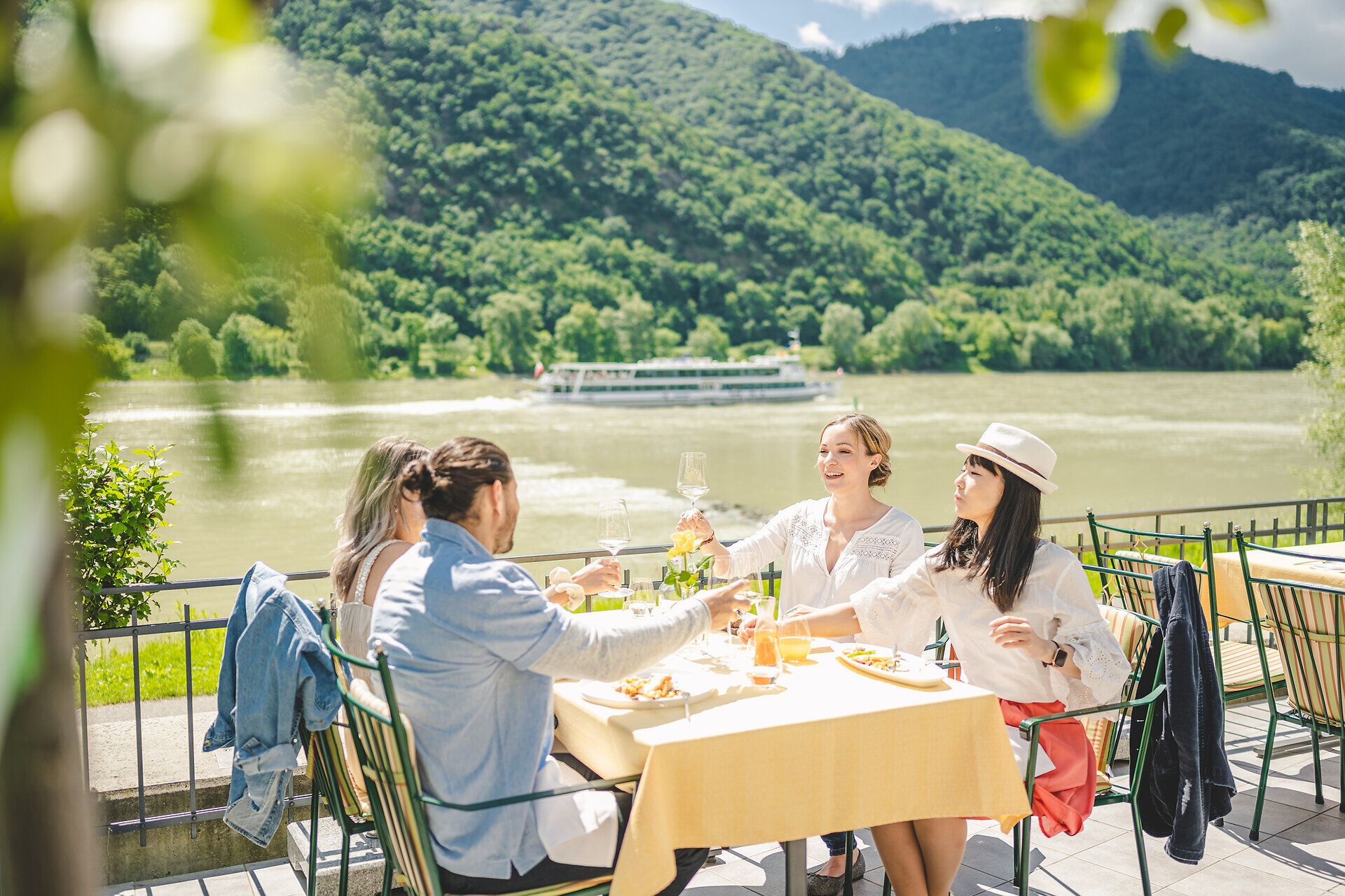 Drei Mädchen und ein junger Bursche sitzen an einem gedeckten Tisch und prosten sich zu.