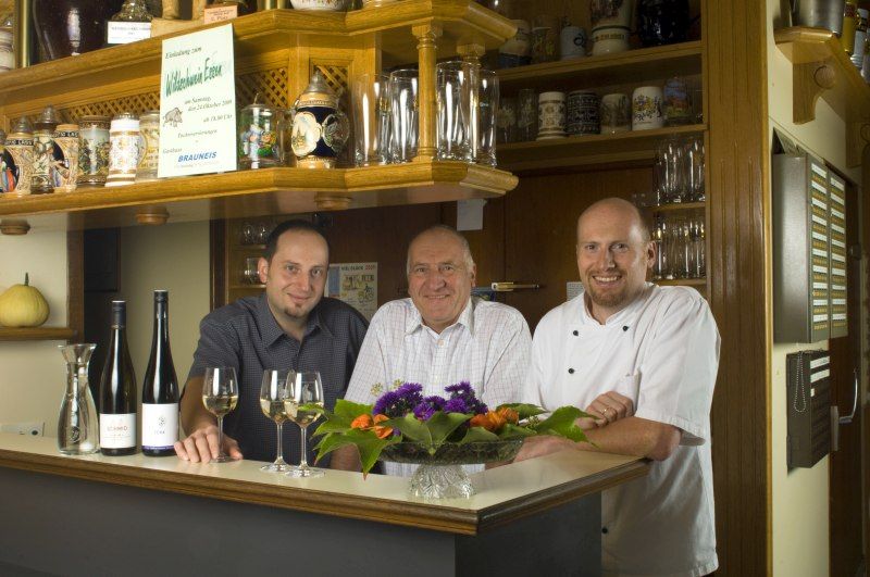 Drei Männer stehen hinter einer Bar mit Weinflaschen und Gläsern.