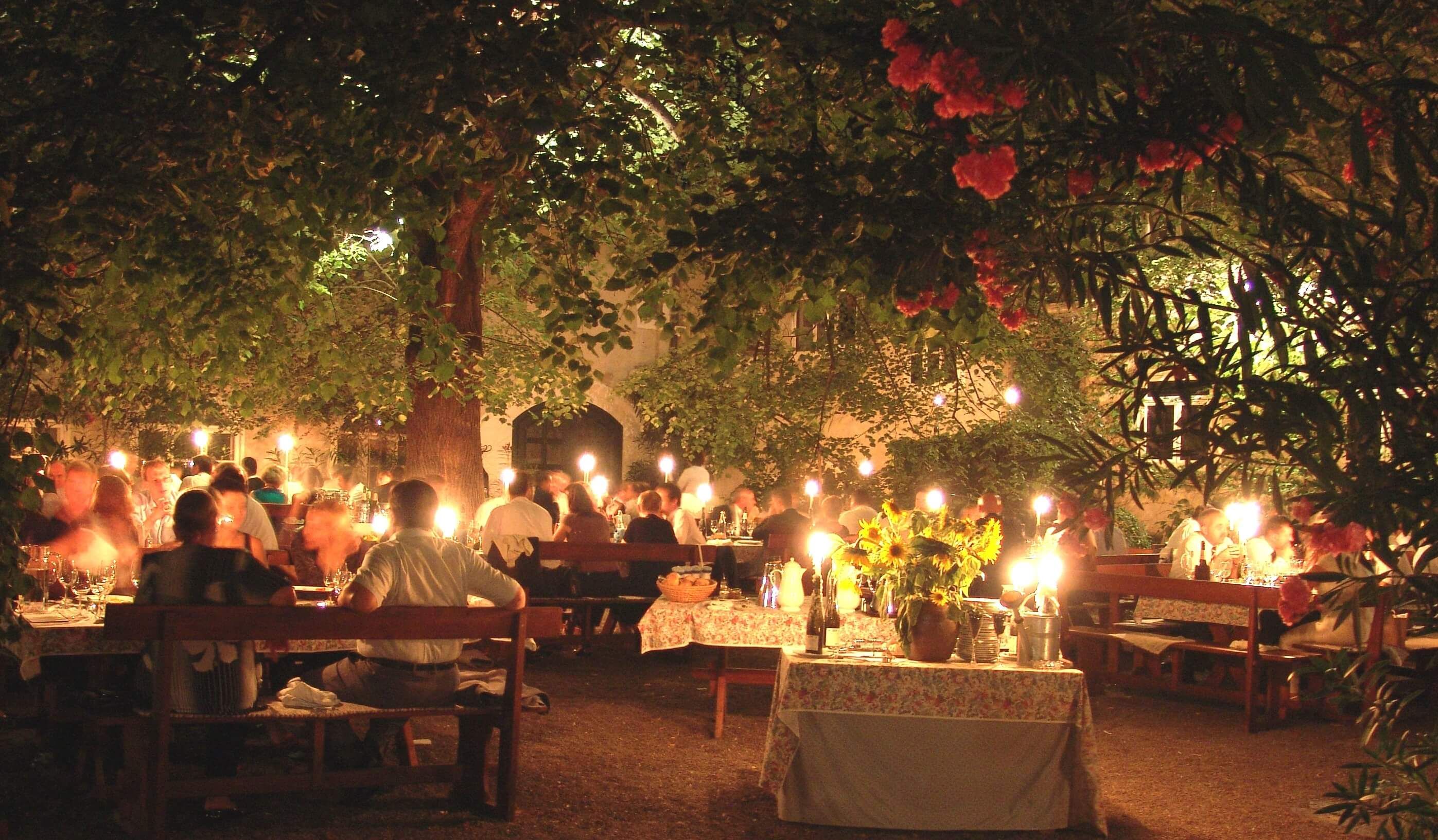 Abendessen im Freien unter einem Baum mit Kerzenlicht und vielen Gästen.