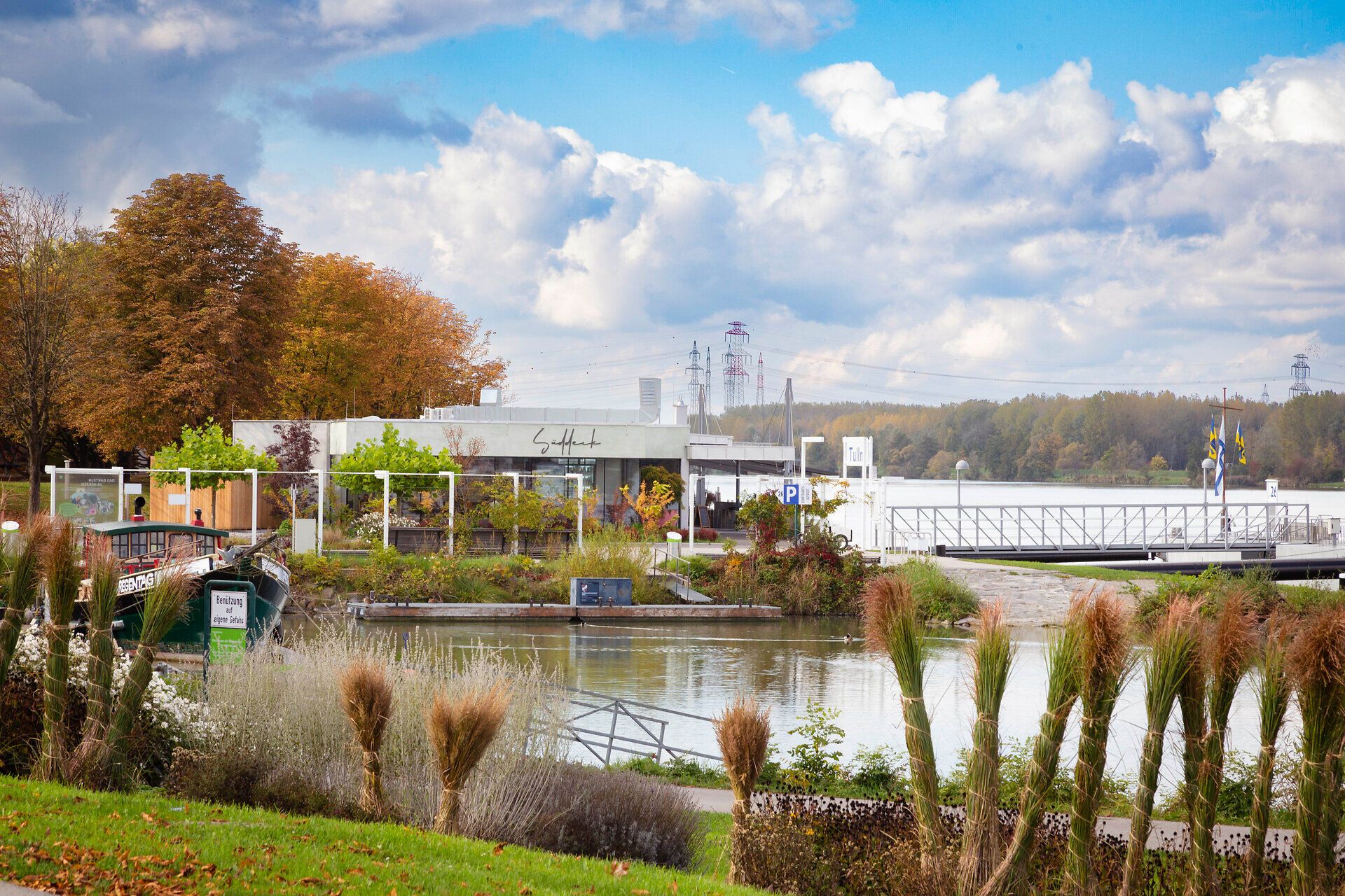 An der malerischen Donaulände entfaltet sich eine harmonische Szenerie, in der sanfte Wellen des Flusses mit dem sanften Licht des Tages verschmelzen. Die bunten Bäume im Hintergrund verleihen der Umgebung eine lebendige Atmosphäre, während die ruhige Wasseroberfläche zum Verweilen einlädt.