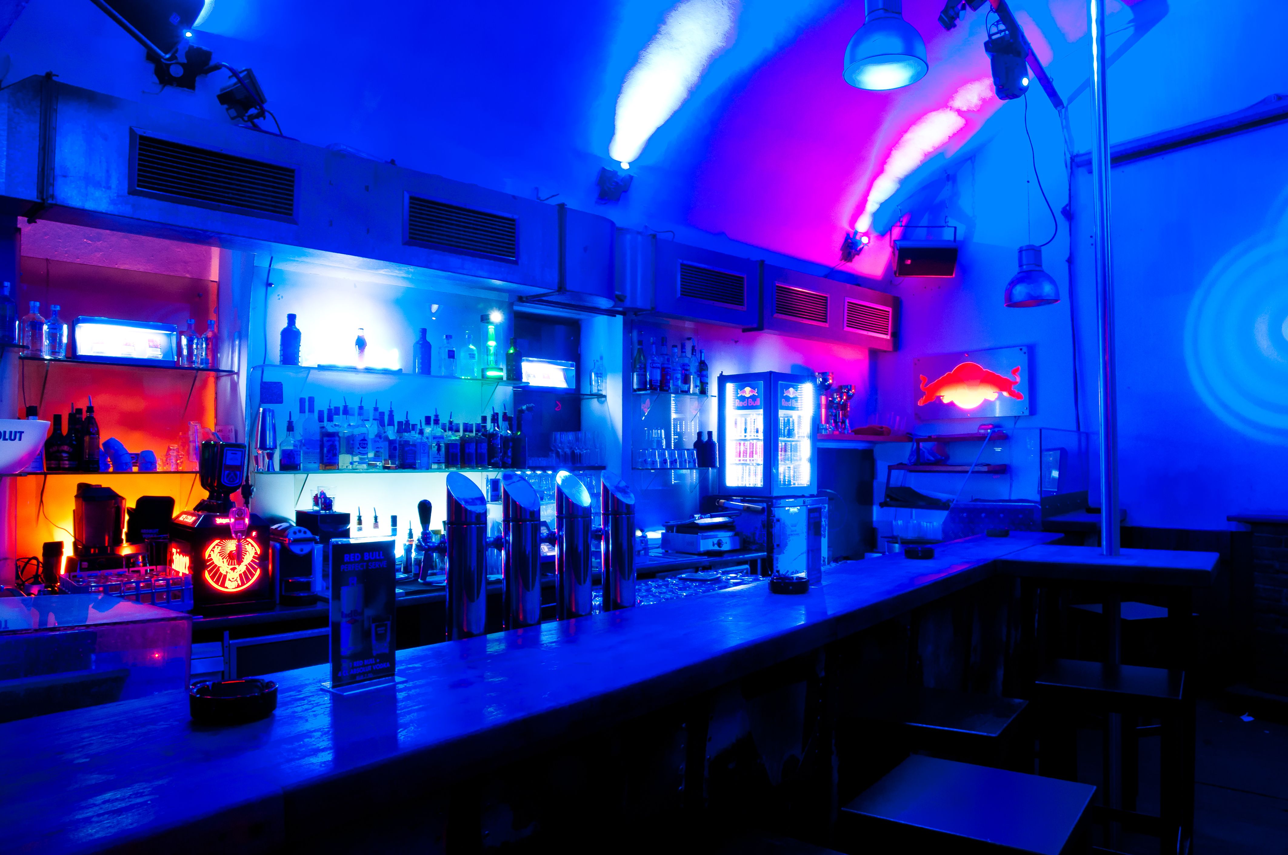 Bar mit blauer und roter Beleuchtung, Flaschen und Gläsern im Regal.
