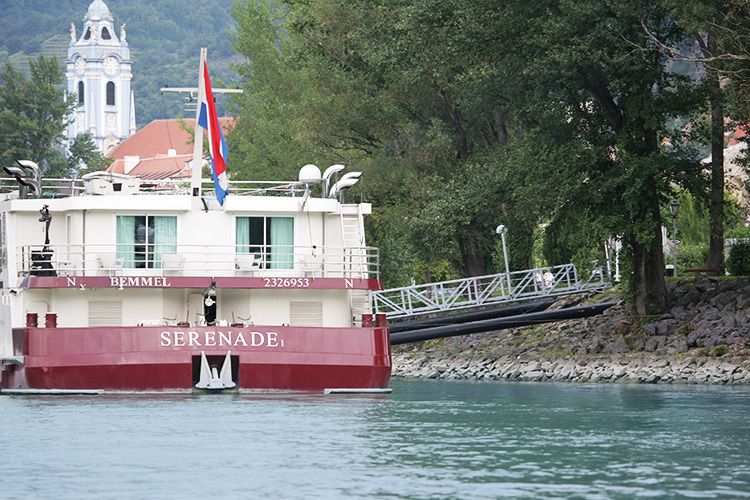Ein Flusskreuzfahrtschiff namens Serenade an einer Anlegestelle in Dürnstein, mit einem Kirchturm im Hintergrund.