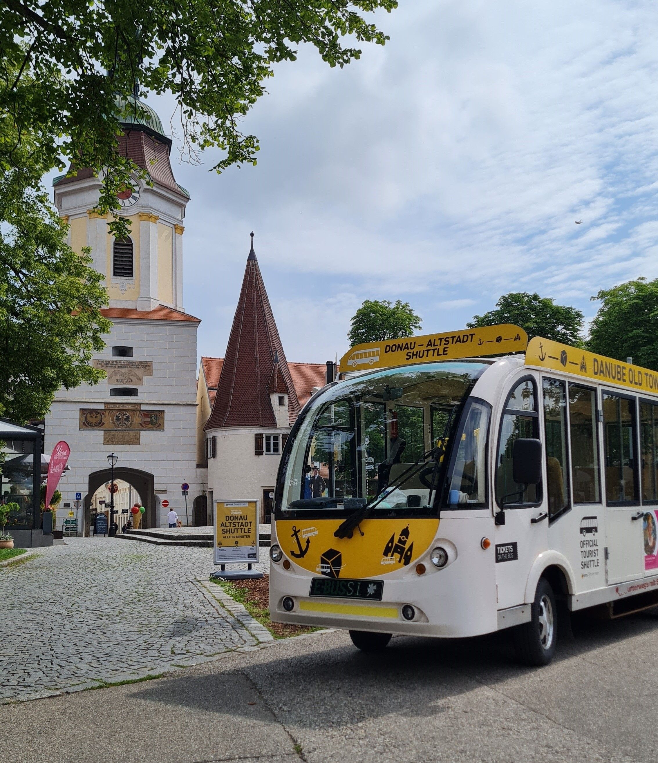 Ein weißer Altstadt-Shuttlebus steht vor einem historischen Torbogen und einem Kirchturm.