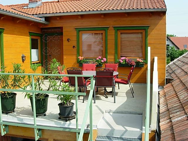 Balkon mit Tisch, Stühlen und Pflanzen, umgeben von einem Haus mit orangefarbener Fassade und grünem Fensterrahmen.