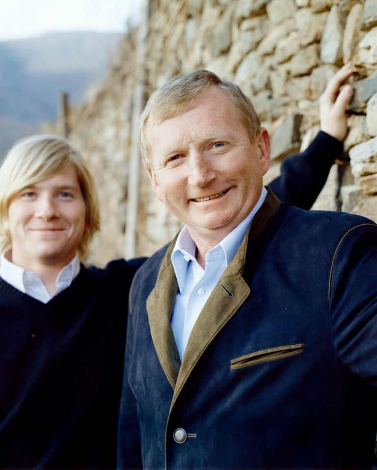 Zwei Männer stehen vor einer Steinmauer, einer lächelt in die Kamera.