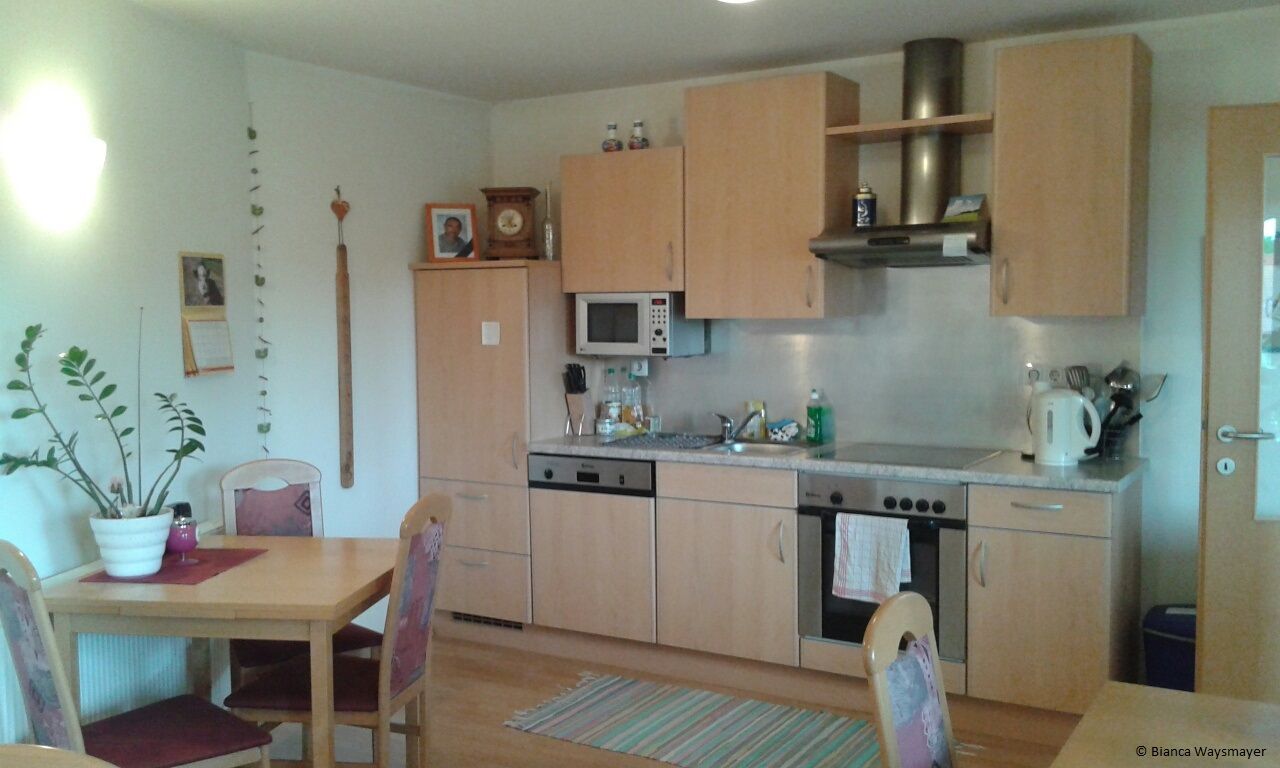 Eine gemütliche Küche mit Holzmöbeln, Esstisch, Stühlen, Spüle, Herd und Mikrowelle. An der Wand hängen Bilder und ein Kalender.