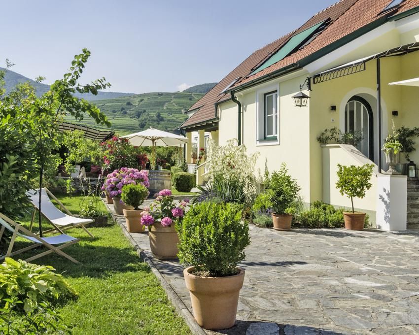 Ein idyllisches Landhaus mit gepflegtem Garten, blühenden Pflanzen und Liegestühlen unter einem sonnigen Himmel.