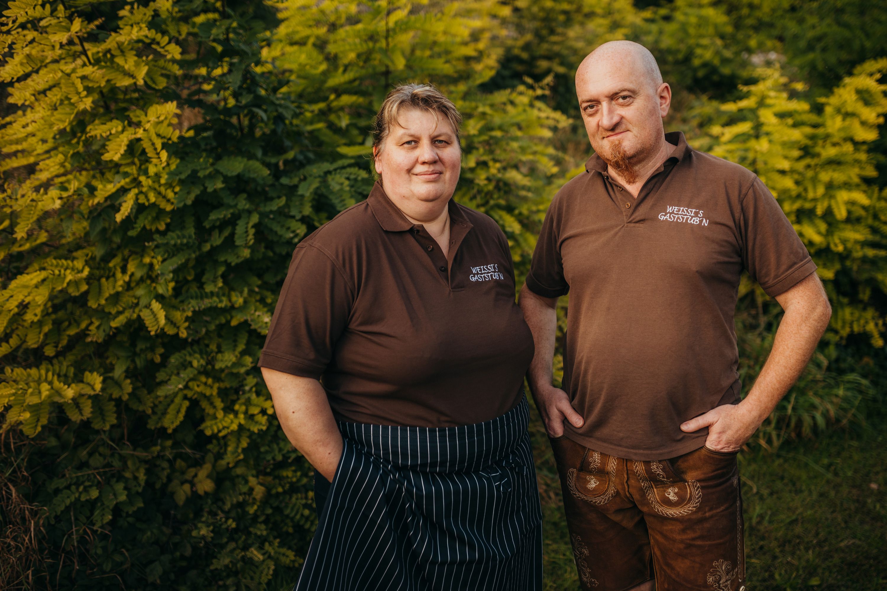 Zwei Personen in braunen Poloshirts vor grünen Büschen.