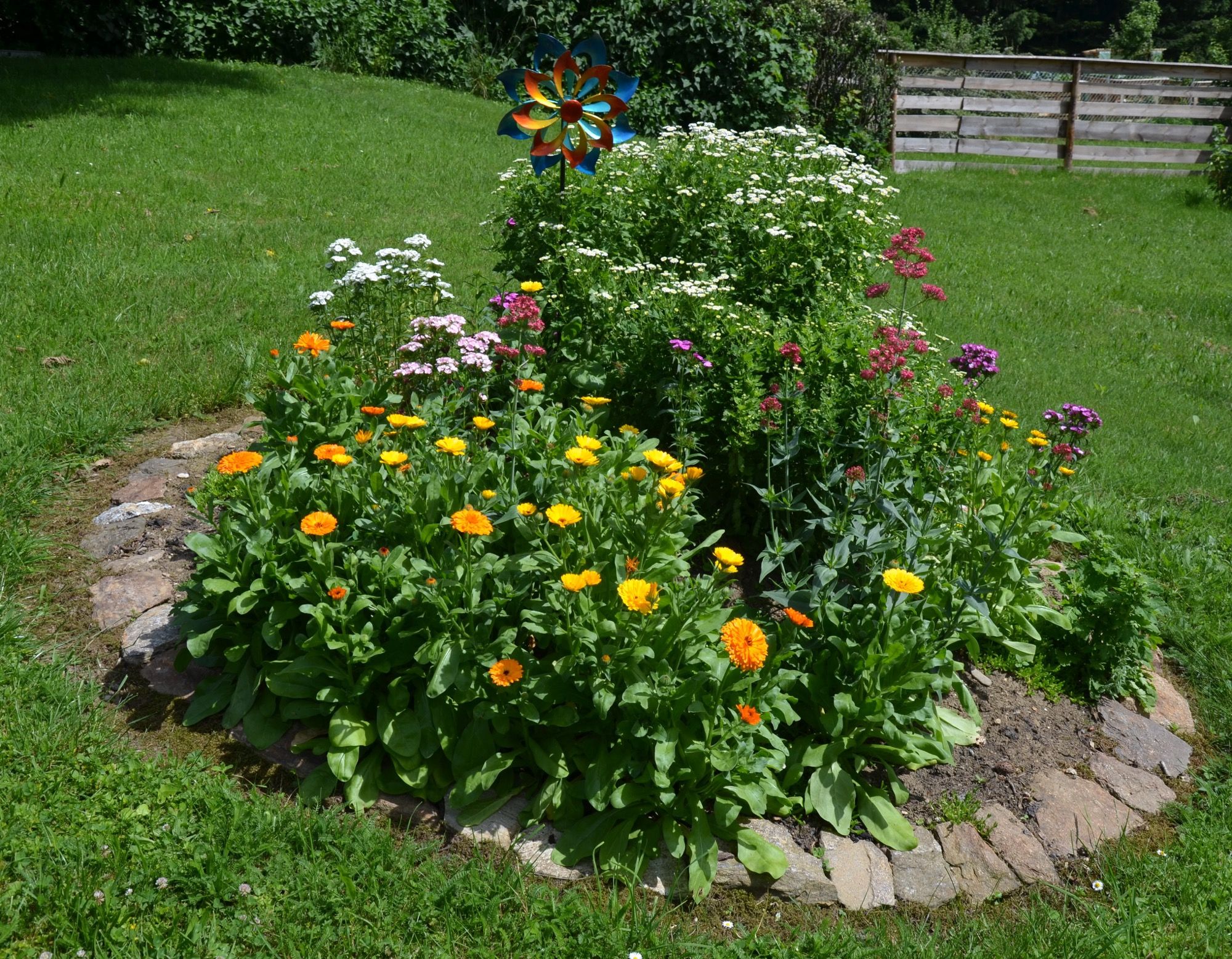 Ein rundes Blumenbeet mit bunten Blumen und einem Windrad im Garten.