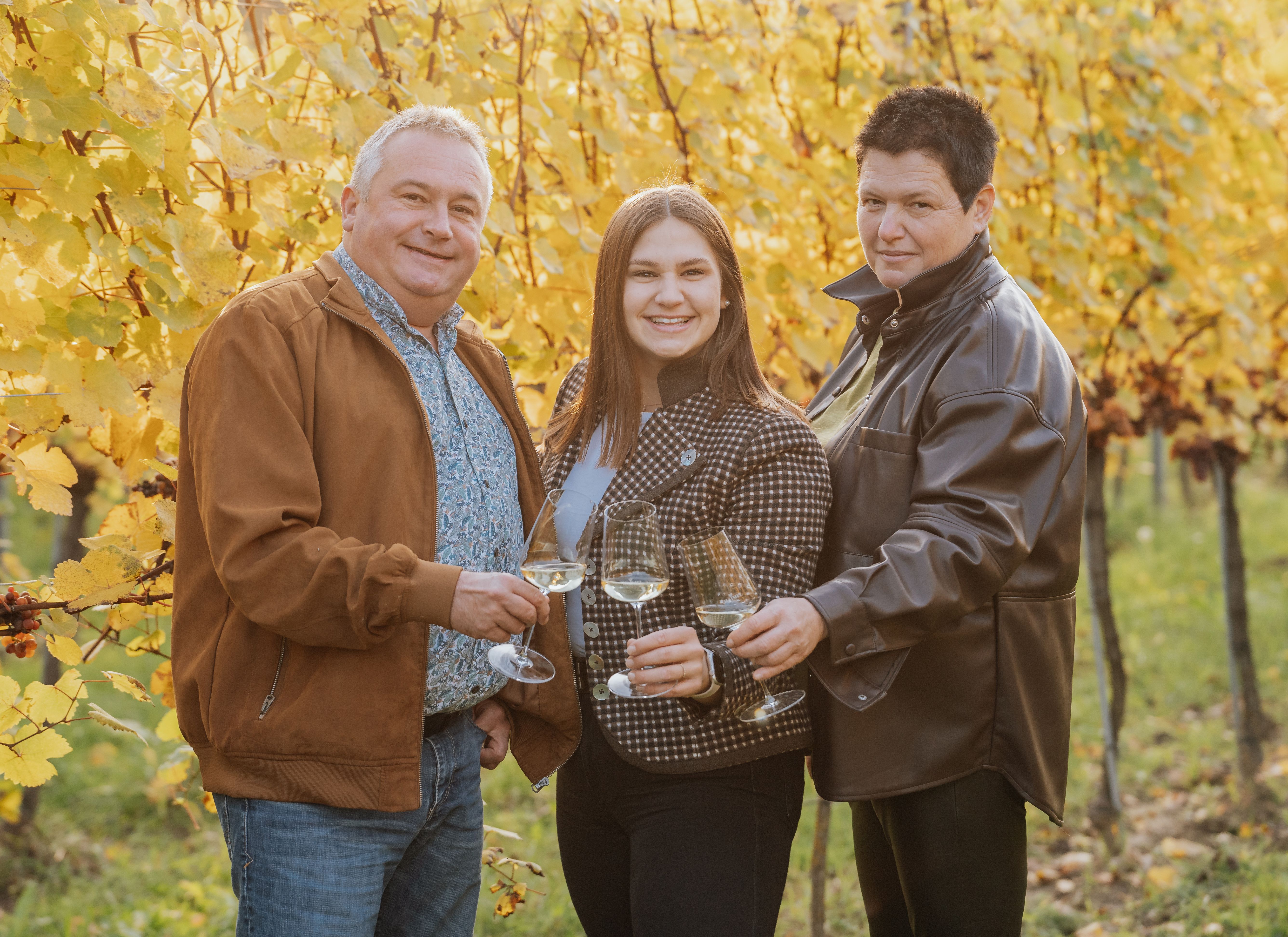 Drei Personen stoßen mit Weingläsern in einem herbstlichen Weinberg an.