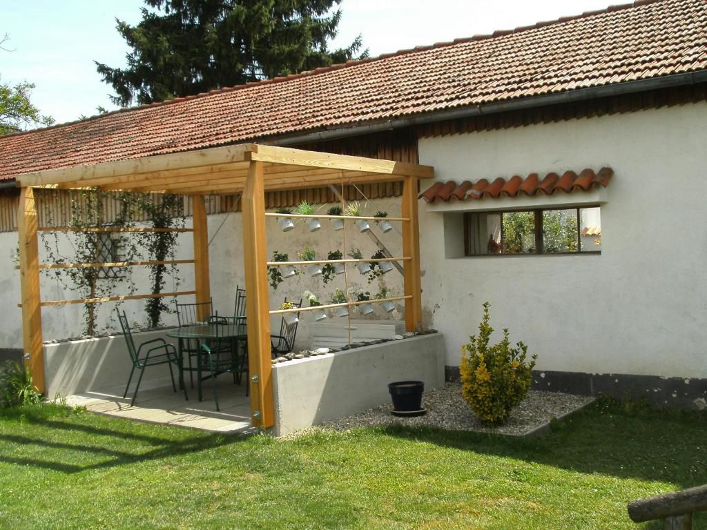 Ein Garten mit einer Holzpergola, darunter ein Tisch und Stühle. Im Hintergrund ein Gebäude mit Ziegeldach.