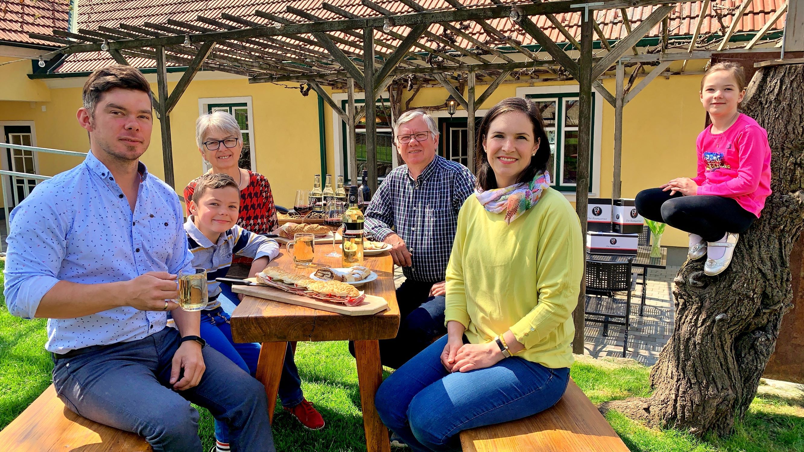 Eine Familie sitzt an einem Holztisch im Freien und genießt eine Mahlzeit. Ein Kind sitzt auf einem Baumstamm. Im Hintergrund ein gelbes Haus mit Pergola.