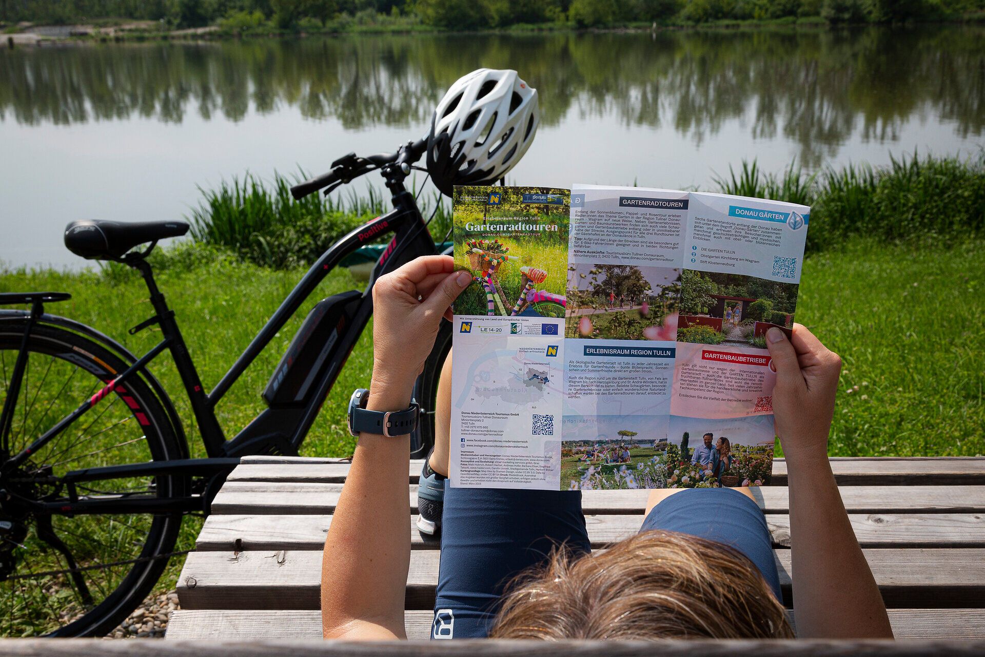 Frau liegt entspannt auf Bank am Wasser und liest Radkarte.