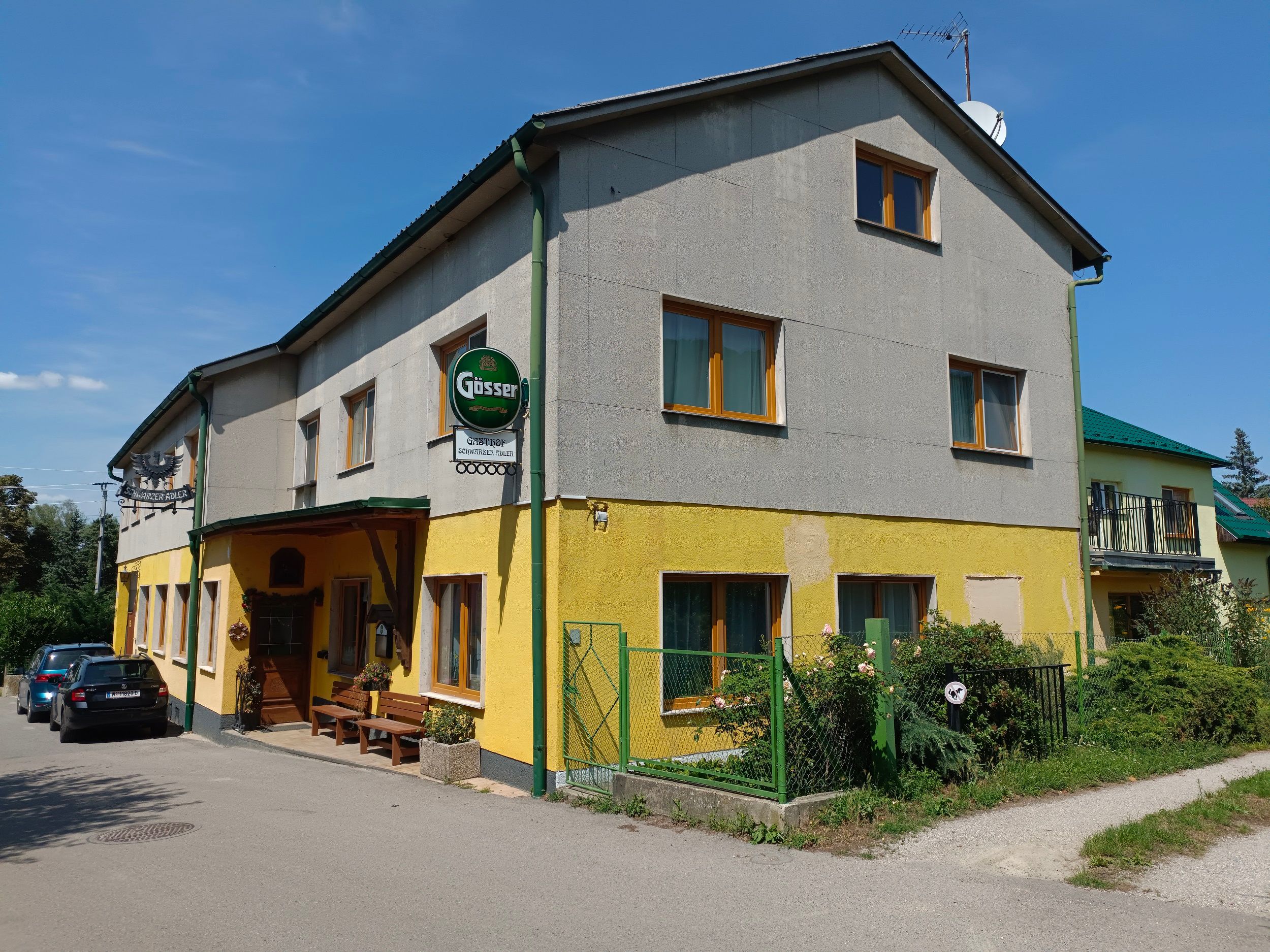 Außenansicht eines gelb-grauen Gasthofs mit Gösserschild und parkenden Autos.