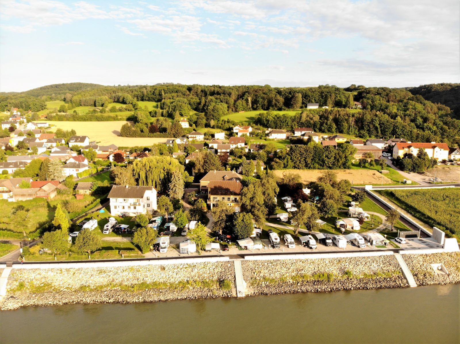 Luftaufnahme eines Campingplatzes am Fluss mit umliegenden Häusern und grüner Landschaft.