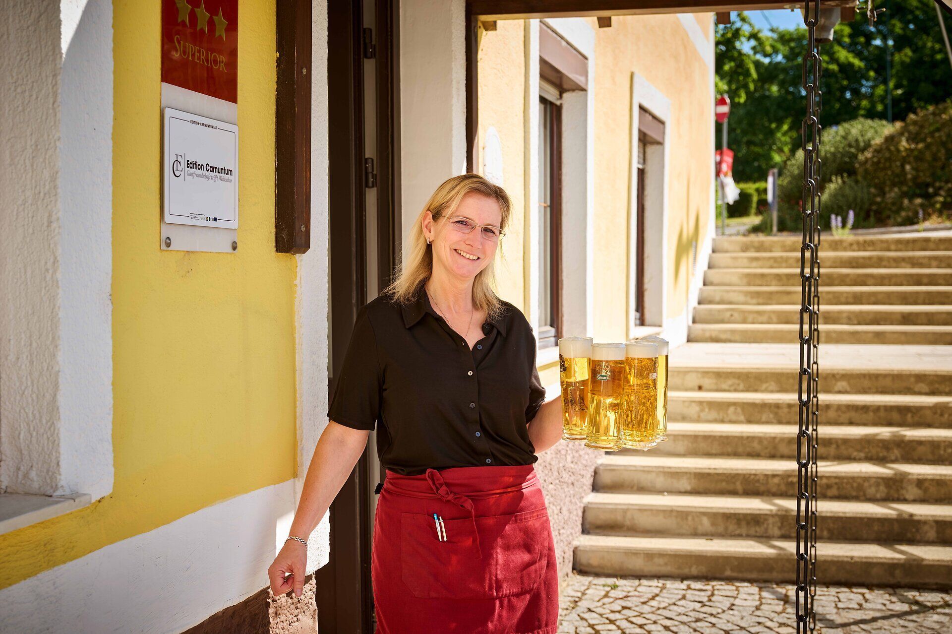 Eine lächelnde Kellnerin trägt vier Gläser Bier vor einem Gasthaus.