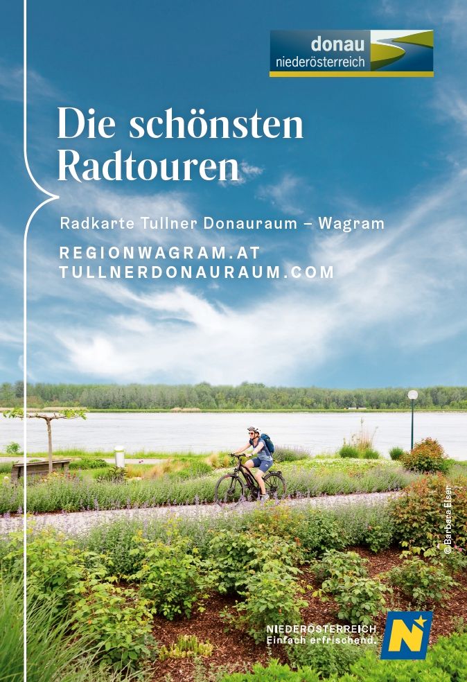 Eine Frau fährt auf einem Radweg in Tulln zwischen schönen grünen Sträuchern und Wiesen. Im Hintergrund sieht man die Donau und einen Auwald.