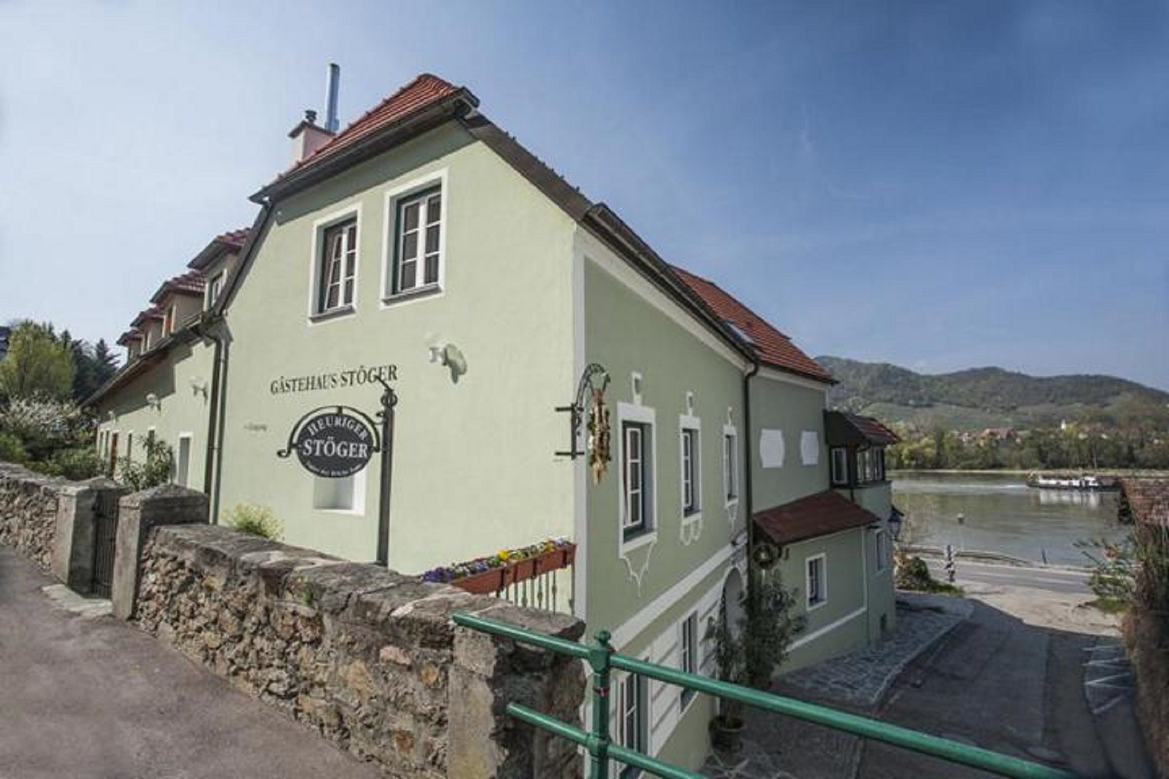 Ein grünes Gästehaus mit roten Dächern am Flussufer, umgeben von Hügeln und blühenden Pflanzen.