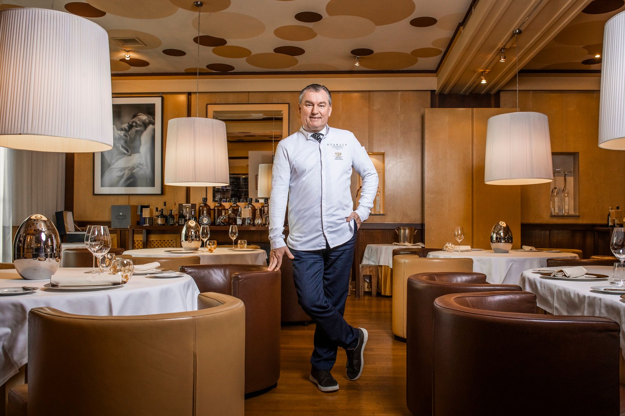 Ein Mann in weißer Kochjacke steht in einem eleganten Restaurant mit runden Tischen und großen Lampen.