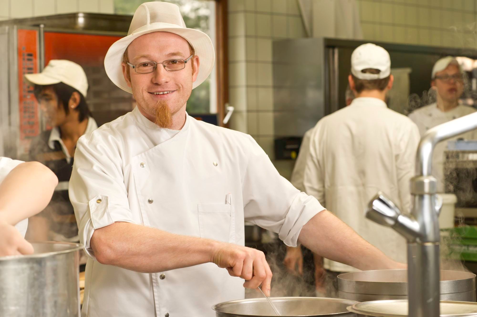 Ein Koch in weißer Uniform und Hut rührt in einem Topf in einer Großküche.