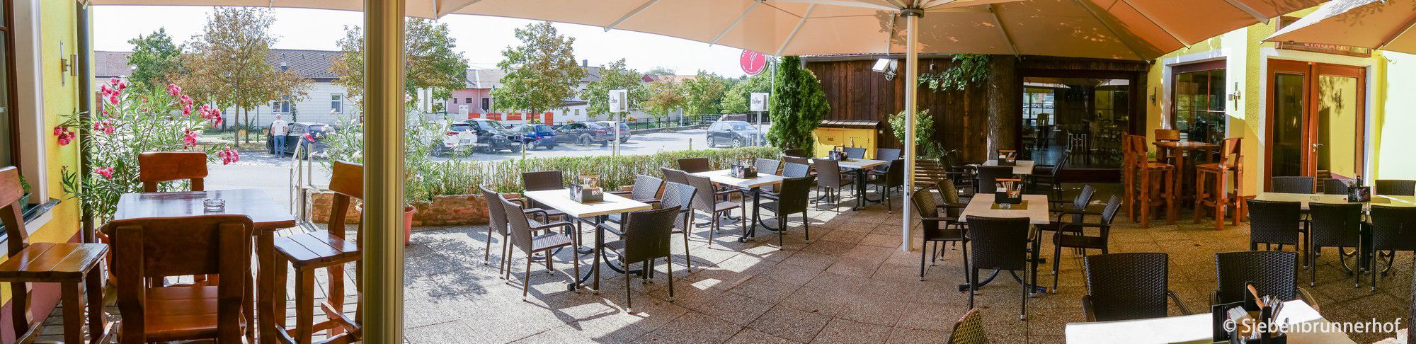 A sunny guest garden with empty tables and chairs, surrounded by plants and a large parasol.