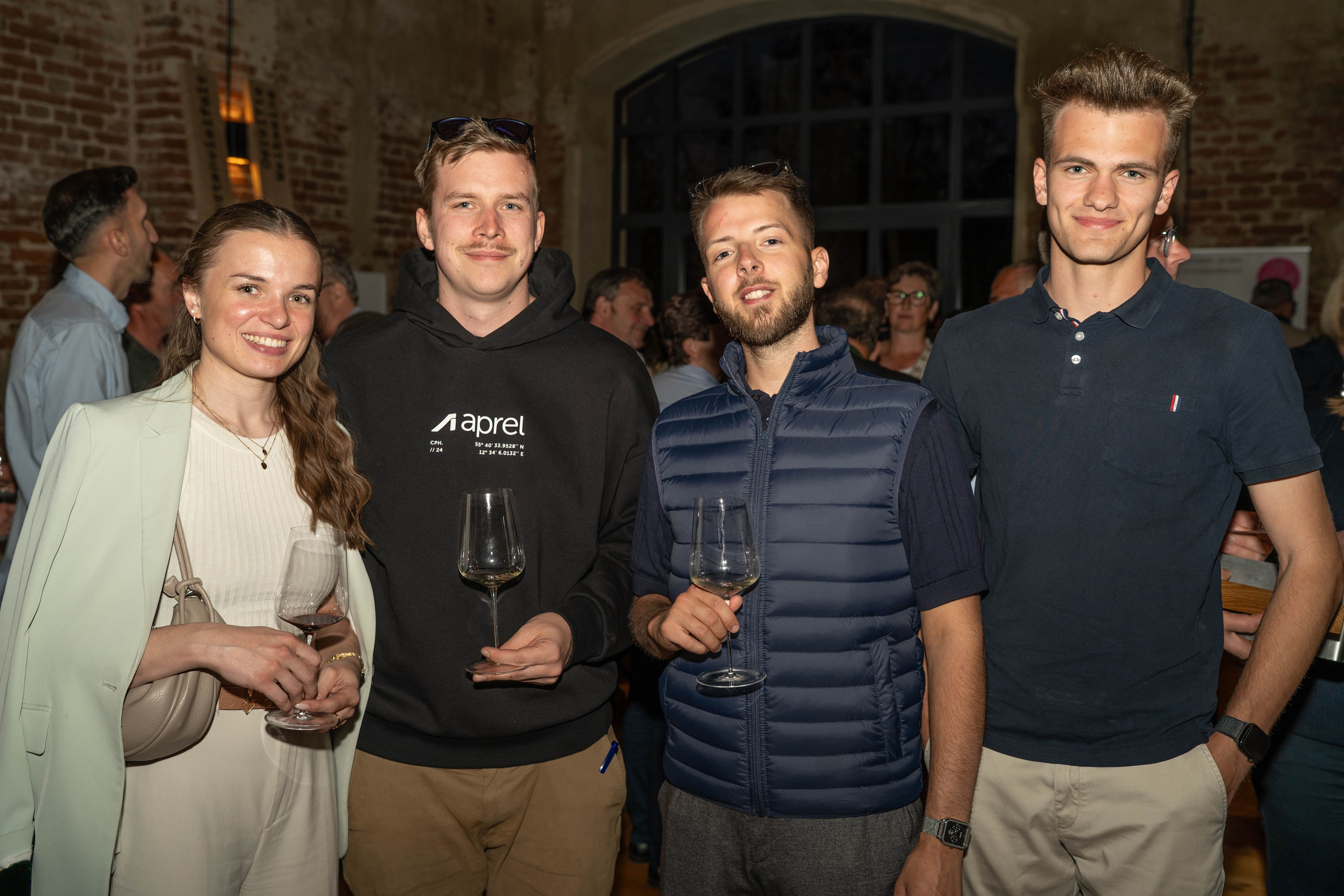 Vier junge Gäste mit Weingläsern bei einer Weinveranstaltung in einer stilvollen Eventlocation mit Backsteinwänden und Abendatmosphäre.