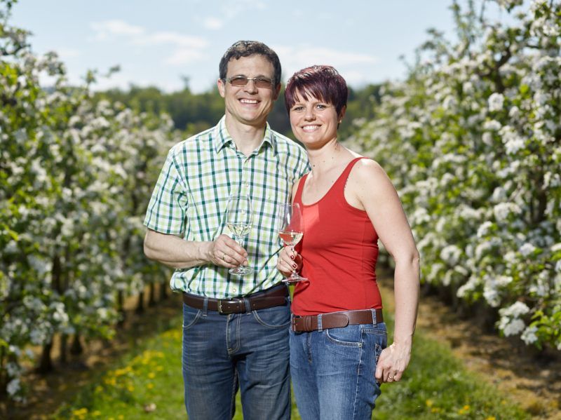 Ein Mann und eine Frau stehen lächelnd in einem blühenden Obstgarten und halten Weingläser.