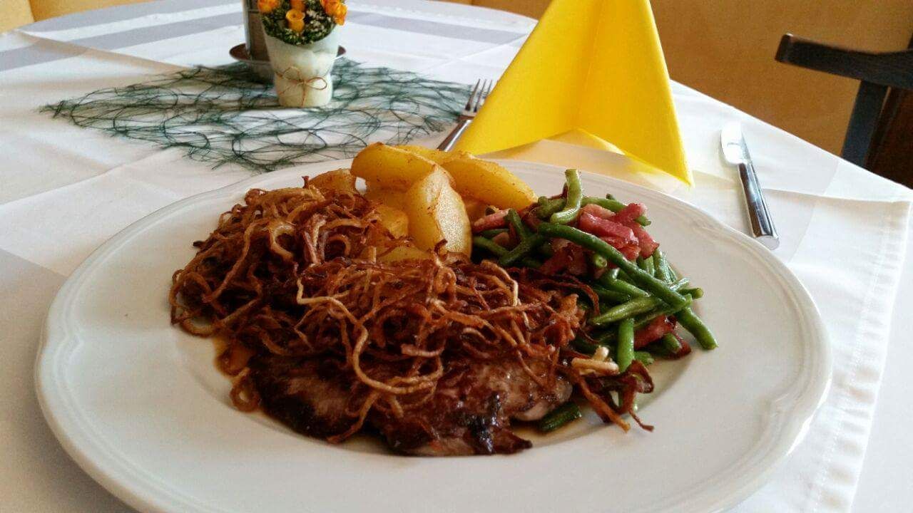 Ein Teller mit Fleisch, Röstzwiebeln, grünen Bohnen und Kartoffeln auf einem Tisch.