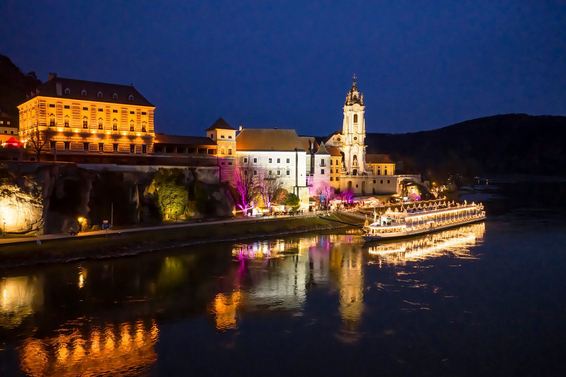 Nächtlicher Blick auf das festlich beleuchtete Dürnstein an der Donau, mit reflektierenden Lichtern, ruhiger Flusslandschaft und historischer Kirche im Advent.
